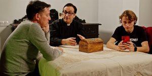 Escena de dos hombres y un joven en una conversación en una mesa, con un sacerdote y un cofre de mad.