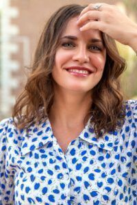 Imagen de una mujer sonriendo, con cabello castaño y vestido azul con lunares blancos, en un entorno.