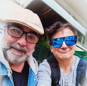Persona con gafas de sol azules y barba, junto a otra con gafas oscuras y mochila.